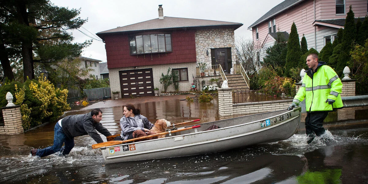 A man in a reflective jacket pulls a boat through a flooded street. Inside the boat, a couple and a dog sit, passing in front of homes surrounded by high water.