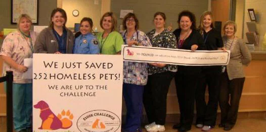 A group of nine smiling people stands indoors, holding two large signs. One sign reads, We just saved 252 homeless pets! We are up to the challenge. The group appears to be celebrating a pet adoption milestone.