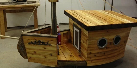 A wooden playhouse designed to look like a small boat, featuring round windows, a small door, and ropes attached to a mast, sitting inside a workshop.