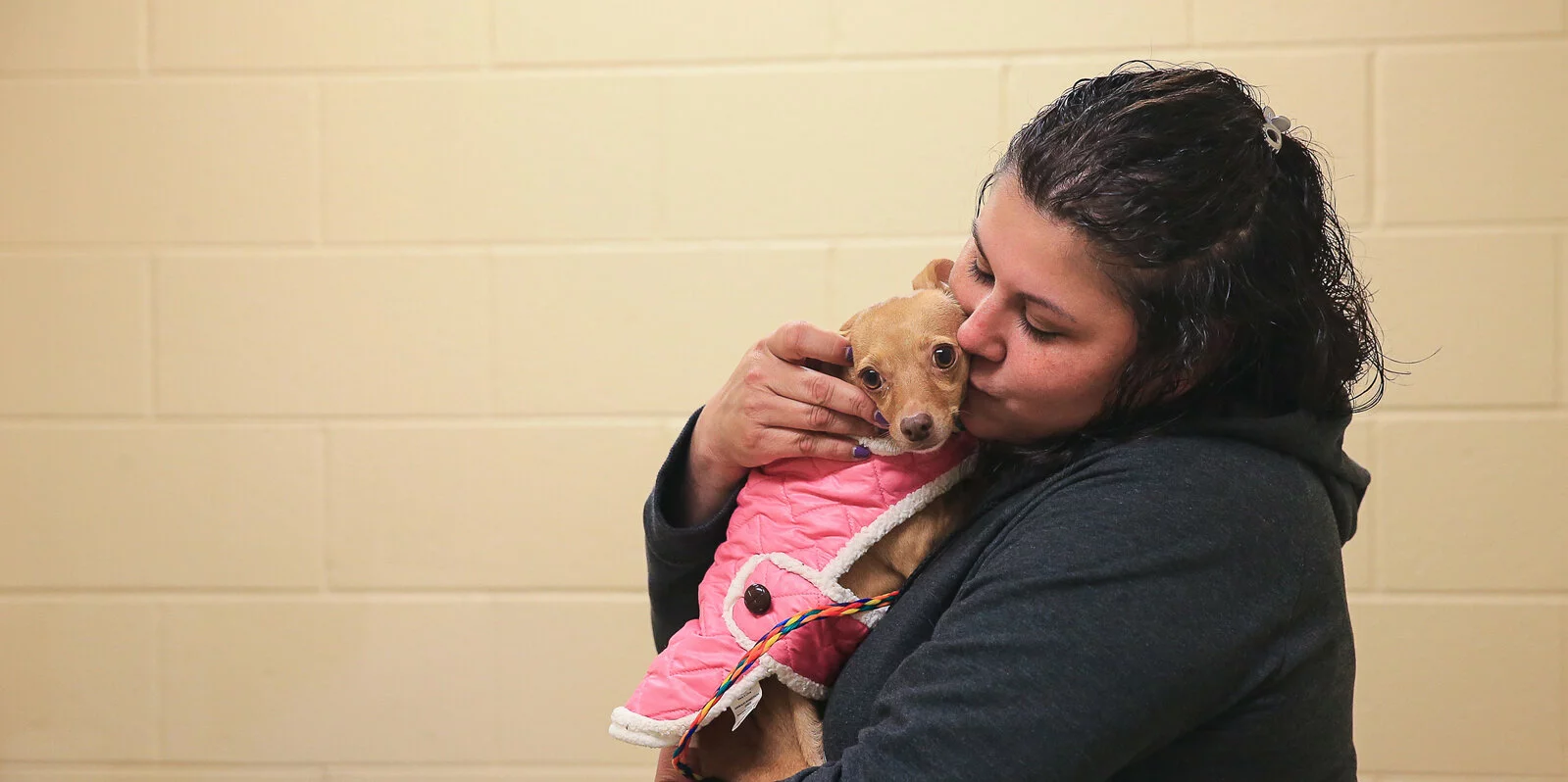 A woman in a dark hoodie hugs and kisses a small tan dog wearing a pink jacket. The dog looks at the camera while the woman shows affection in front of a beige brick wall.