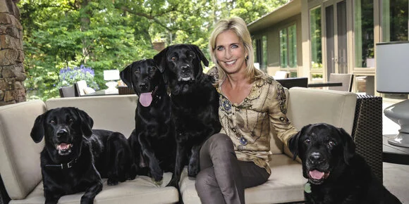 A woman sits on an outdoor couch smiling, surrounded by four black Labrador dogs, with greenery and a modern house in the background.