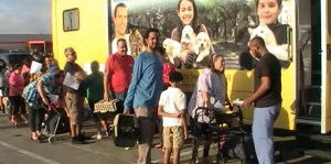A diverse group of people with pets wait in line outside a yellow mobile clinic. Some hold carriers or paperwork, while a staff member assists an elderly woman near the entrance. A mural of smiling people with animals is on the vehicle.