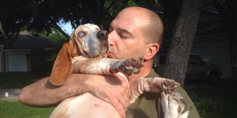 A man gently kisses a brown-and-white dog while holding it in his arms outdoors, with trees, a house, and a vehicle in the background.