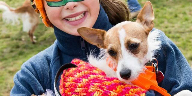 A smiling child wearing a colorful mask and hat holds a small dog dressed in a bright knitted sweater outdoors on a grassy field.