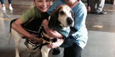 Two children kneel and smile while hugging a large dog with brown and white fur indoors. The dog wears a harness and leash. The background is blurred, showing more people and a concrete floor.