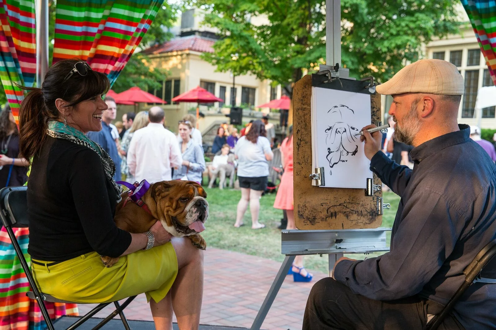 A woman sits with a bulldog on her lap while an artist sketches the dog at an outdoor event with people mingling and colorful decorations in the background.