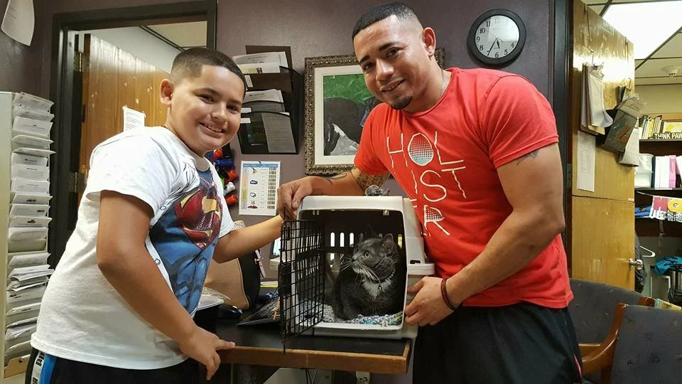 A smiling man and boy stand beside a table, holding a pet carrier with a gray cat inside, in what appears to be a veterinary office. The boy wears a Superman shirt, and the man wears a red Hollister shirt.