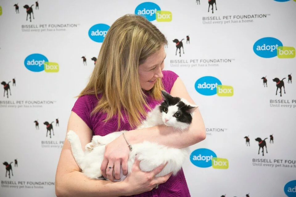 A woman in a pink top holds a black and white cat in her arms, standing in front of a backdrop with adoptbox logos and Bissell Pet Foundation branding.