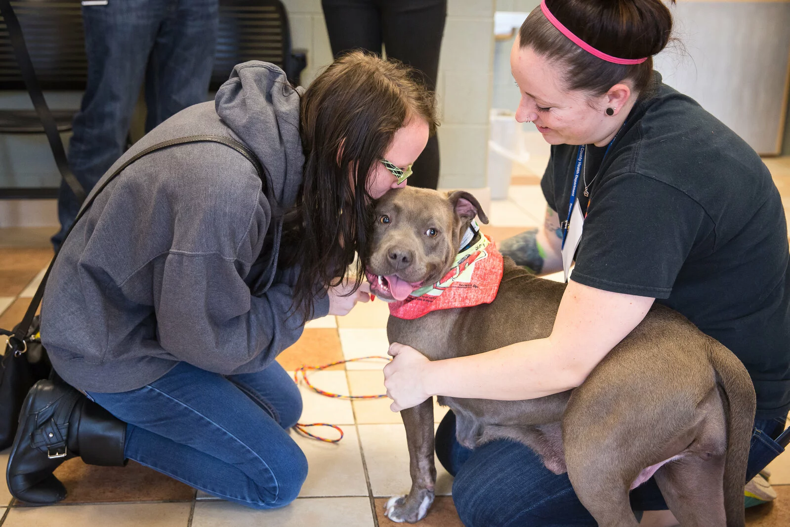 Two women kneel beside a gray pit bull wearing a red bandana; one woman smiles while hugging the dog, and the other leans in close, while the dog looks at the camera, tongue out, in a tiled indoor setting.