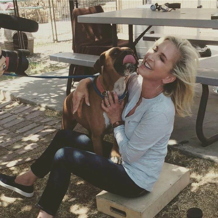 A woman sitting on a wooden box by a picnic table smiles and laughs as a brown dog on a leash licks her face. She is wearing a light blue shirt and jeans, and the scene appears to be outdoors on a sunny day.