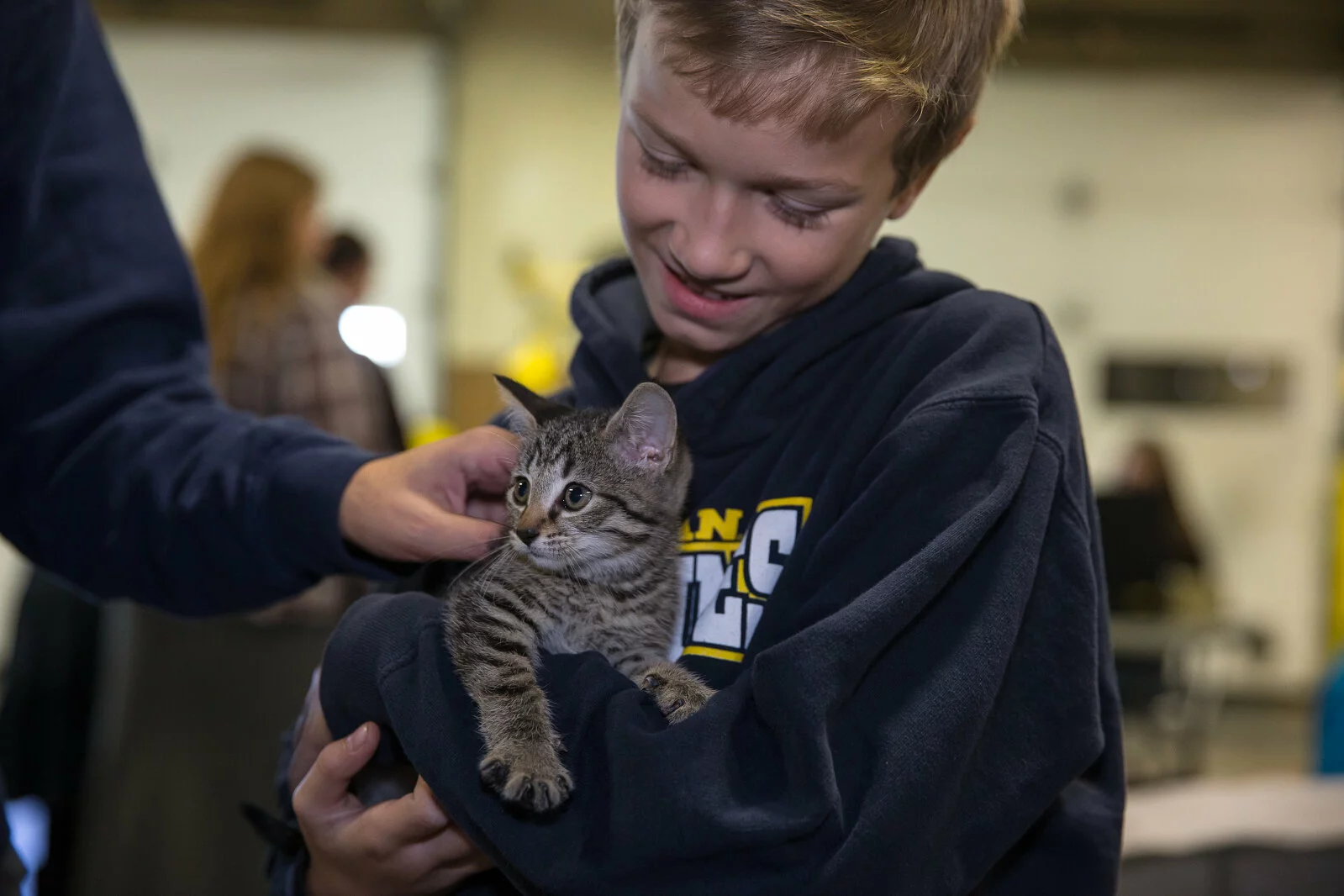 A smiling boy in a navy hoodie gently holds a gray tabby kitten while another person pets the kitten’s head. The background is softly blurred indoors, with a few people visible.