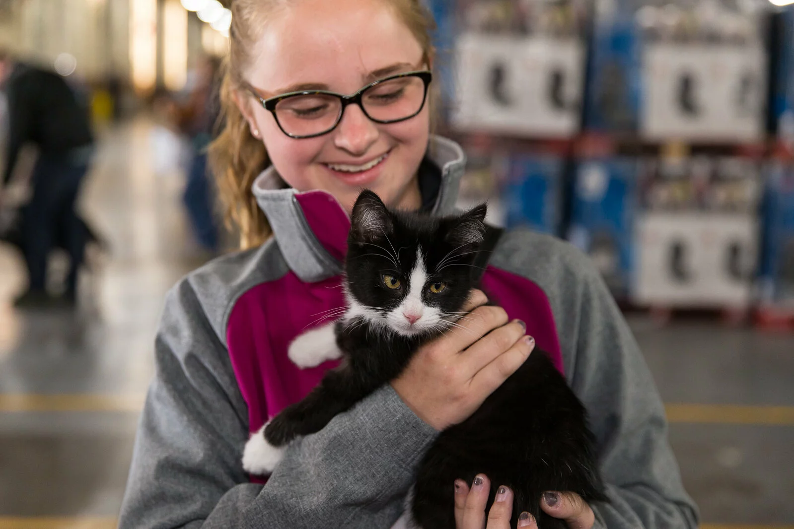 A smiling person with glasses and a gray and magenta jacket holds a black and white kitten indoors, with shelves and people blurred in the background.