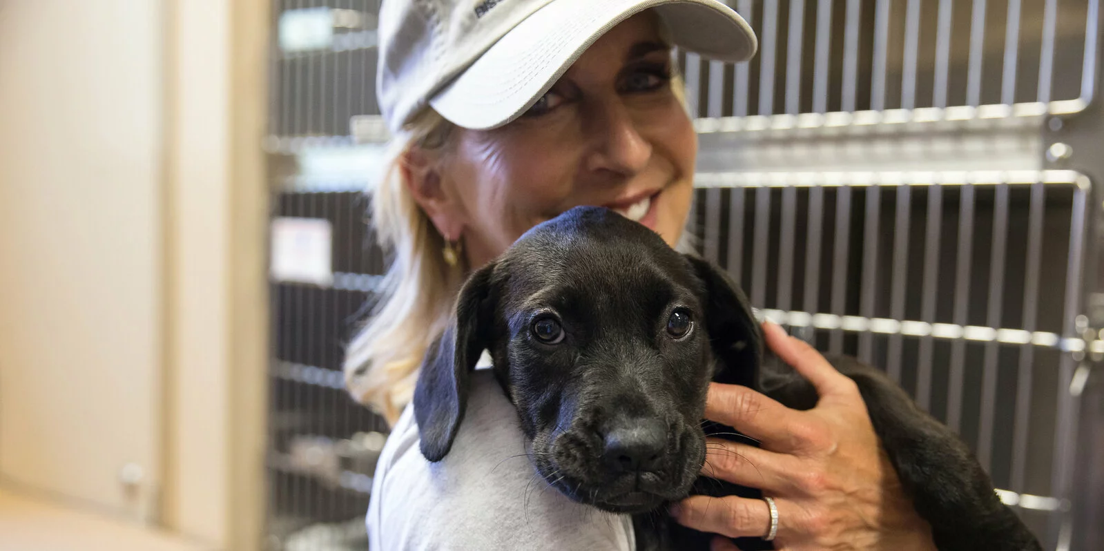 A smiling woman wearing a cap holds a black puppy close to her chest inside an animal shelter, with kennel cages visible in the background.
