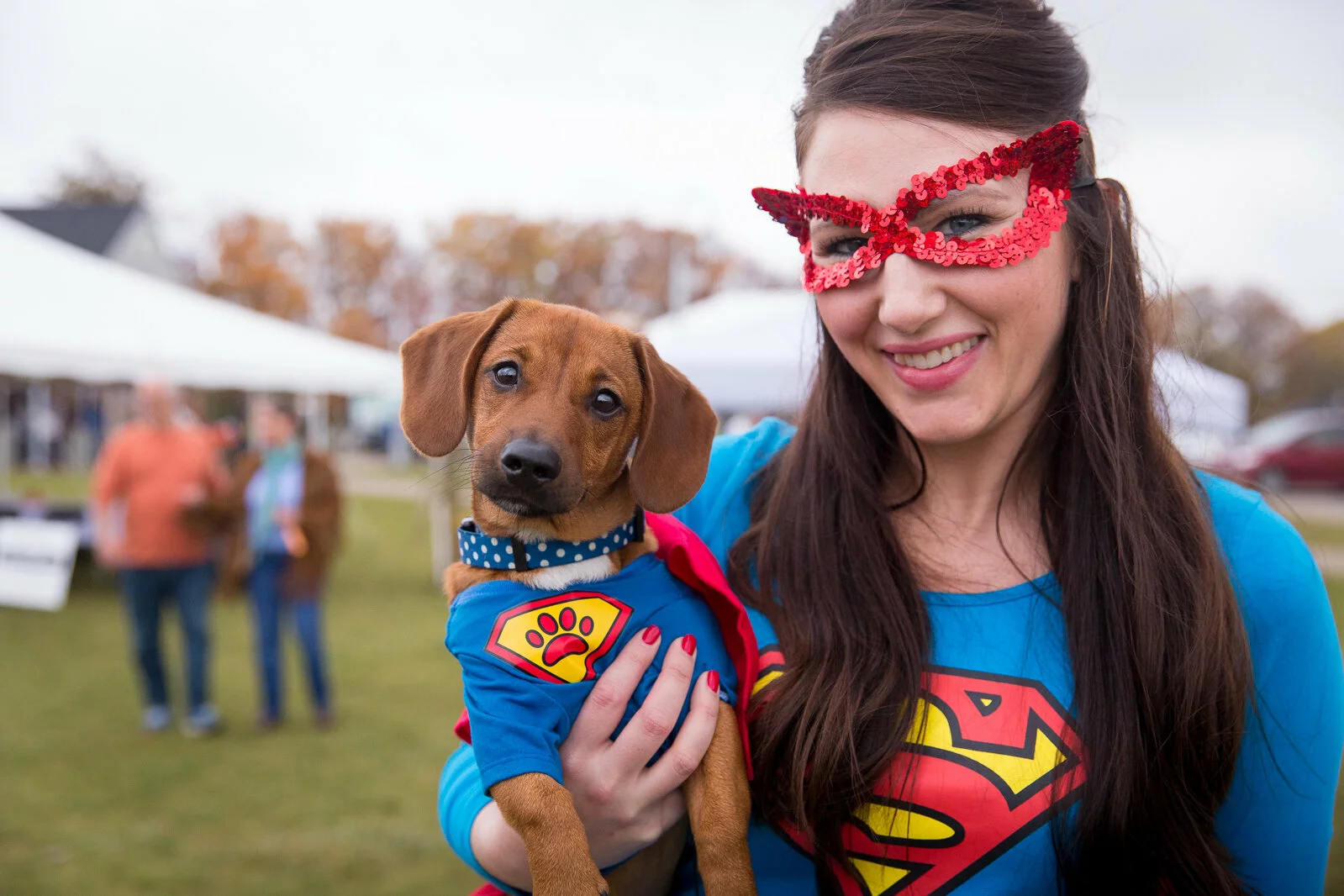 A smiling woman in a red glittery mask and Superman shirt holds a brown puppy dressed in a blue cape and superhero shirt at an outdoor event. White tents and people are visible in the background.