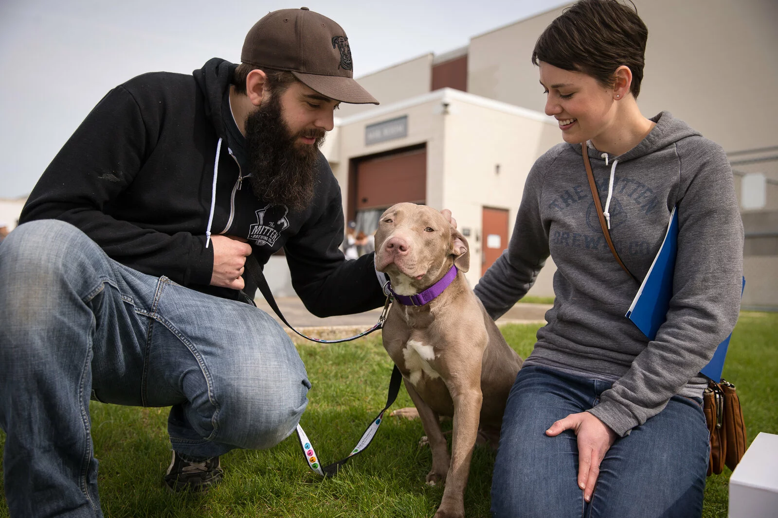 A man and woman kneel on grass, smiling and petting a gray pit bull wearing a purple collar. The man holds the dog’s leash. There is a building with garage doors in the background.