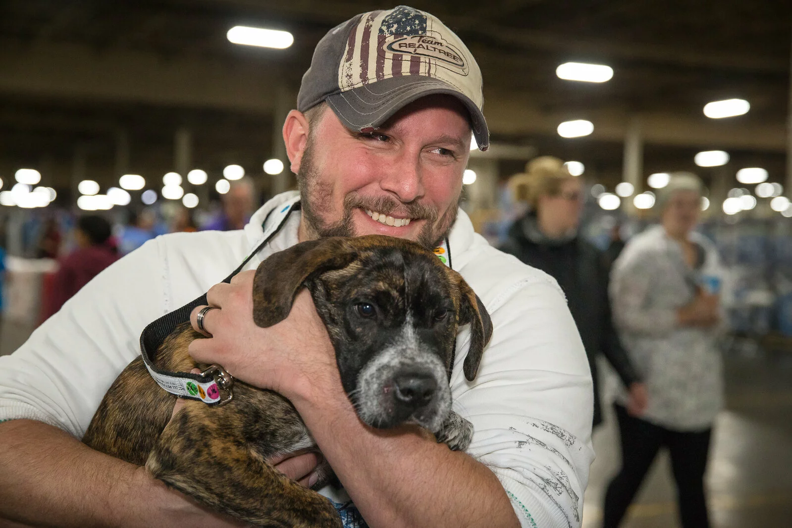A smiling man in a baseball cap hugs a brindle dog closely indoors, with people and lights blurred in the background.