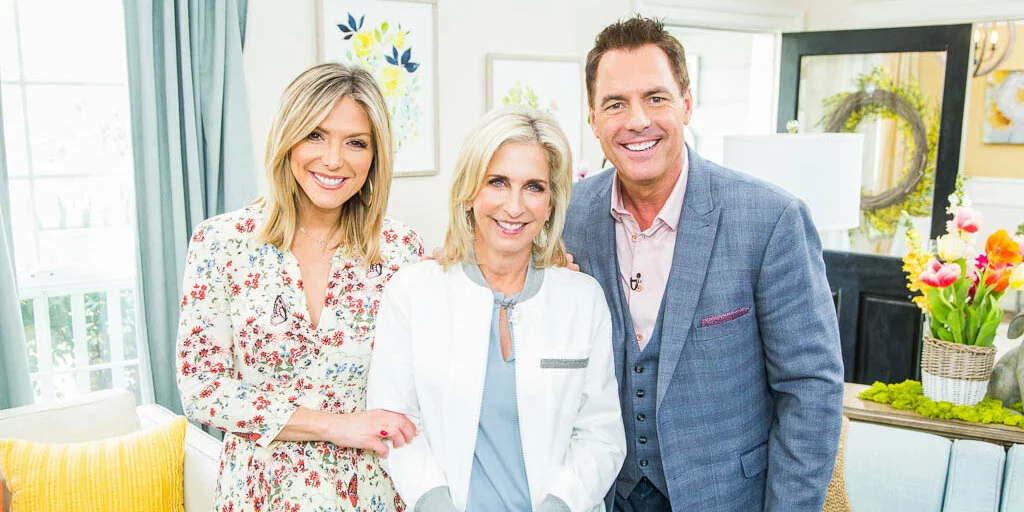 Three people are smiling indoors, standing close together in a brightly lit, decorated living room with floral accents and pastel colors. Two women and one man are dressed in business-casual attire.