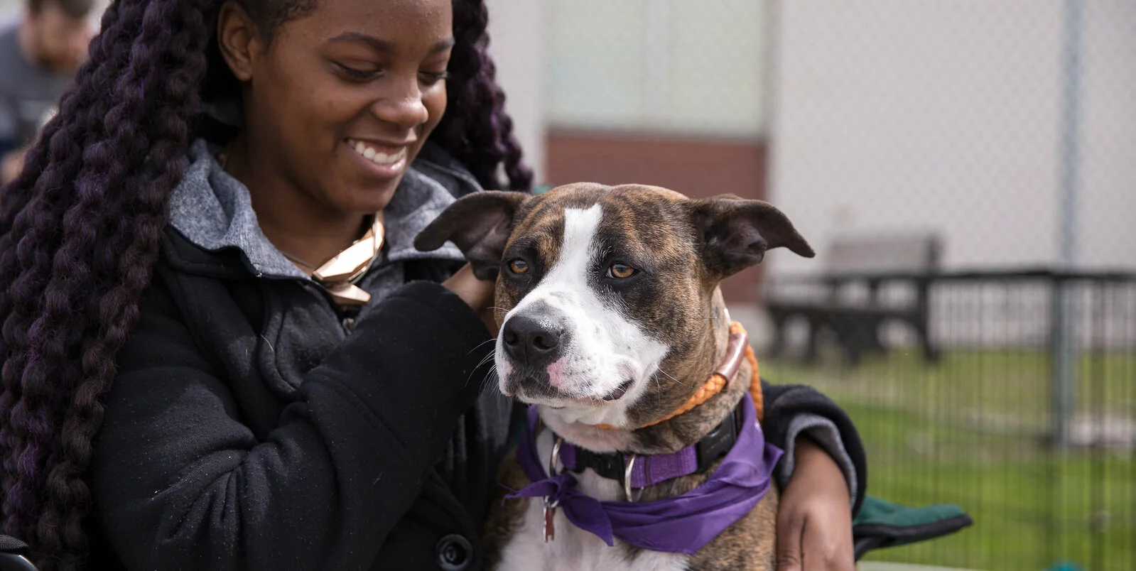 A woman with long, braided hair smiles while petting a brown and white dog wearing a purple bandana and orange collar, outdoors near a fence and benches.