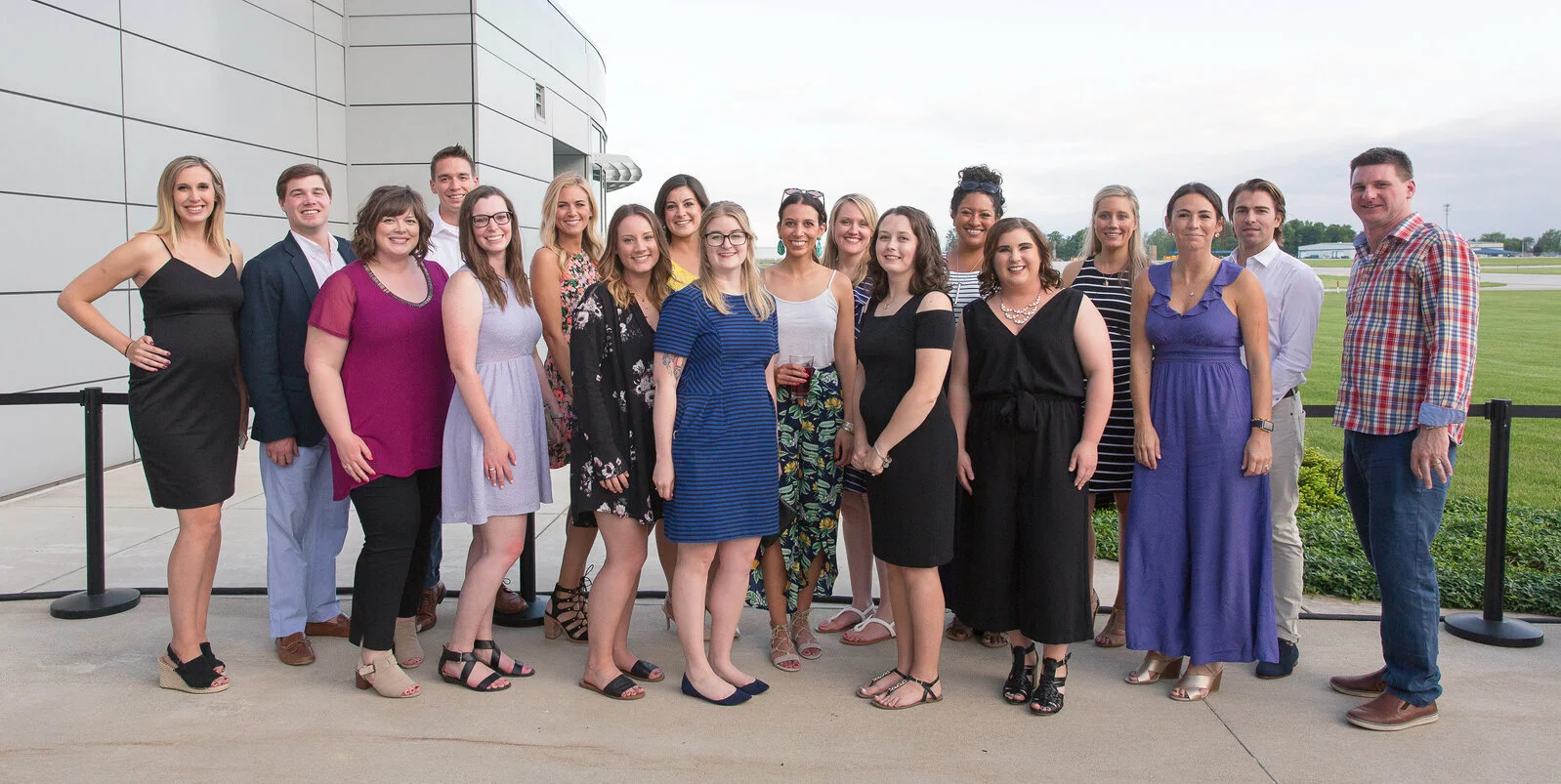 A group of 17 people stand together outside a modern building, smiling at the camera. They are dressed in semi-formal and casual attire, with greenery and an open field visible in the background.