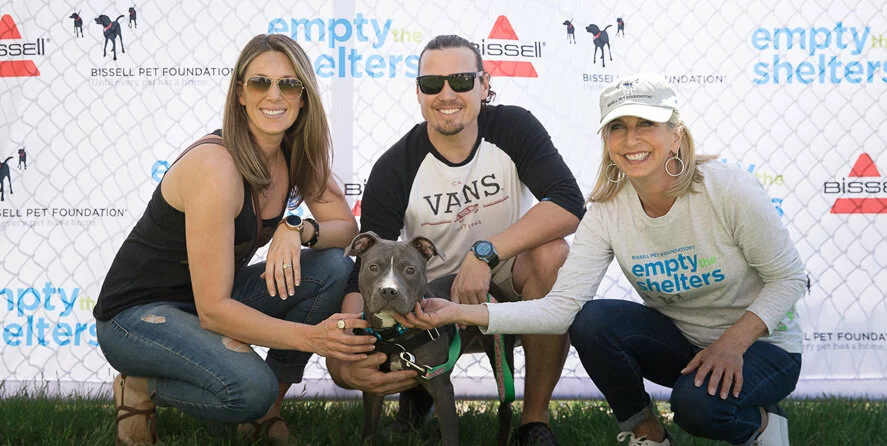 Three smiling adults pose with a gray dog in front of a fence with “Empty the Shelters” and “Bissell Pet Foundation” logos, suggesting a pet adoption event.