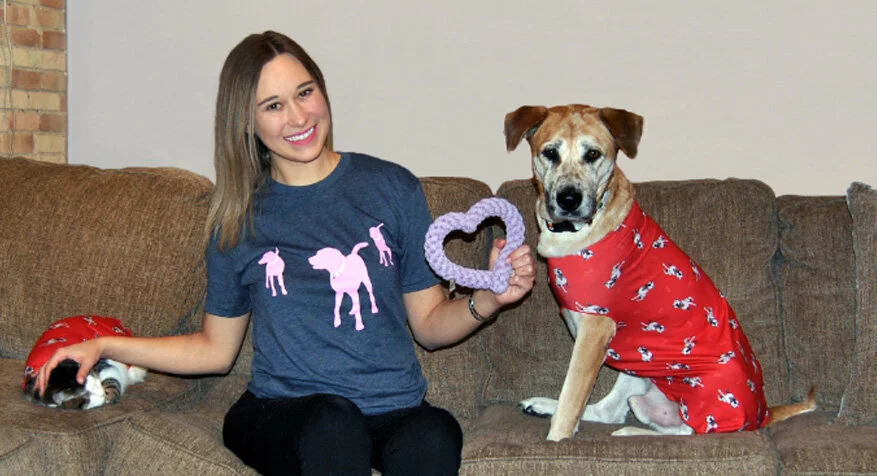 A woman smiles while sitting on a couch, wearing a blue shirt with pink dog silhouettes. Next to her is a dog in red pajamas holding a purple heart-shaped toy, and a cat in matching red pajamas lying at her side.