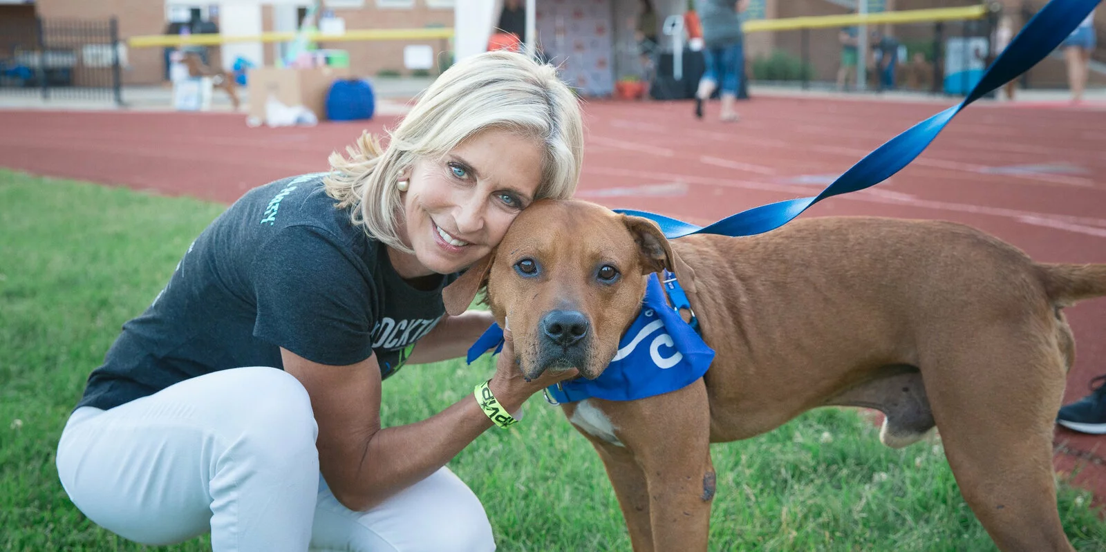 A smiling woman with blonde hair kneels on grass next to a brown dog wearing a blue bandana. The woman hugs the dog, and they both face the camera on an outdoor track field.