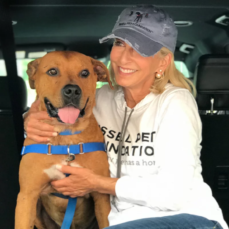A smiling woman wearing a cap and white sweatshirt sits in a car, hugging a happy brown dog with a blue harness.