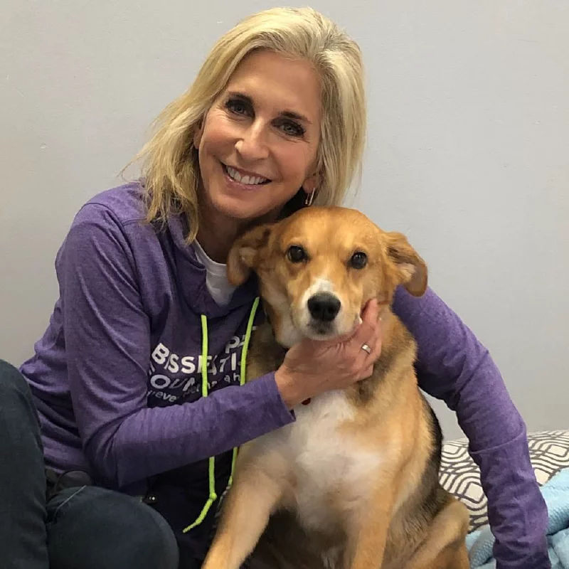 A smiling woman with blonde hair, wearing a purple hoodie, sits indoors and gently hugs a brown and white dog while posing for the camera.