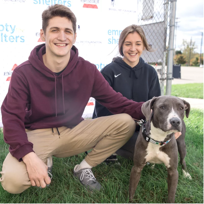 Two smiling people kneel on grass with a gray and white dog, posing for a photo in front of a fence and a backdrop with Empty the Shelters logos. The dog wears a black collar and looks toward the camera.