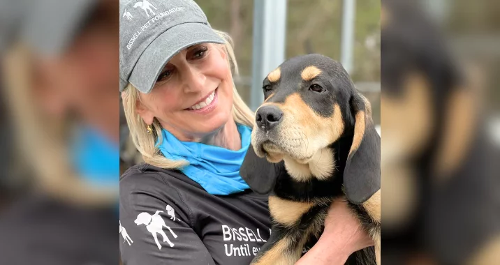 A smiling woman wearing a gray cap and black shirt holds a large black and tan puppy outdoors. The woman appears happy, and the puppy looks calm.