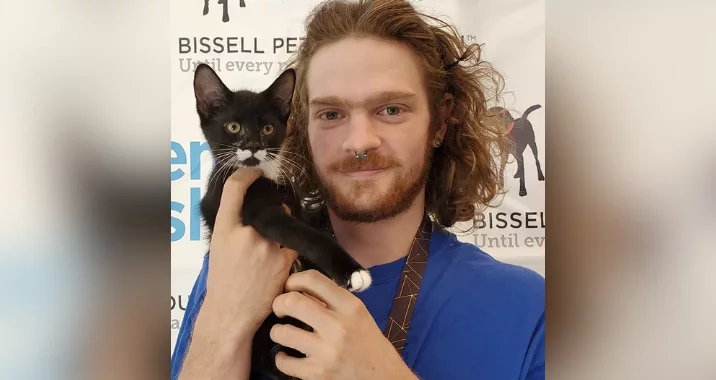 A person with long curly hair and a nose ring smiles while holding a black and white kitten in front of a Bissell Pet Foundation backdrop.