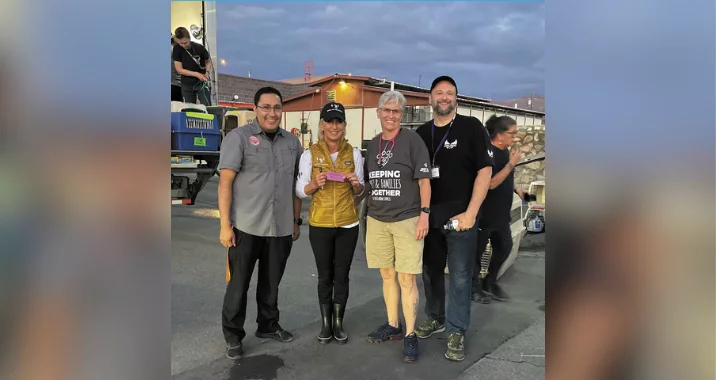 Four adults stand smiling outdoors in casual clothing at dusk, with a utility vehicle and building in the background. Two people are holding an object together while others stand beside them.