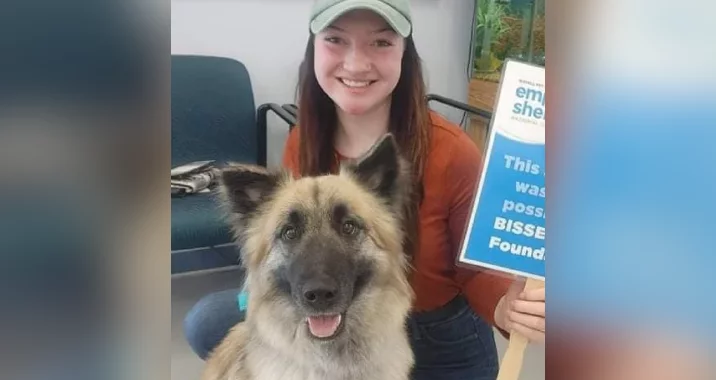 A smiling woman in a green cap and orange shirt sits indoors next to a happy, fluffy dog. She holds a sign with blue text, partially visible, promoting Empty the Shelters in what appears to be an animal shelter setting.