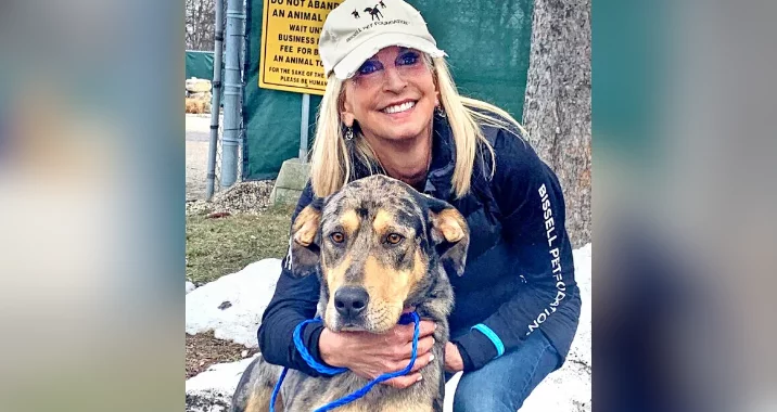 A smiling woman in a beige cap and dark jacket crouches outdoors, hugging a large, brown and black dog with a blue leash—enjoying the joy of an Empty the Shelters event. Trees and a yellow sign are visible in the background.