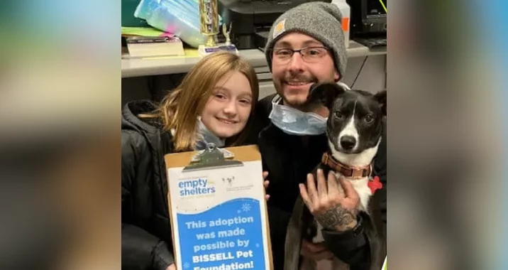 A smiling man and young girl pose with a black and white dog. The man holds a sign reading, This adoption was made possible by BISSELL Pet Foundation’s Empty the Shelters! They appear happy and are indoors.