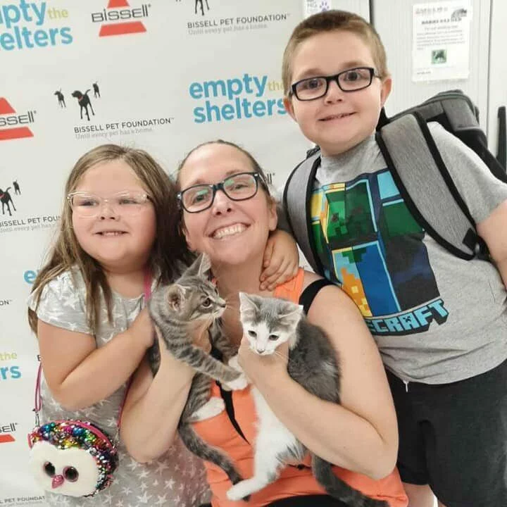 A smiling woman and two kids, both in glasses, pose with kittens in front of an Empty the Shelters sign. The girl sports a sequined owl purse as they each hold or pet a kitten.
