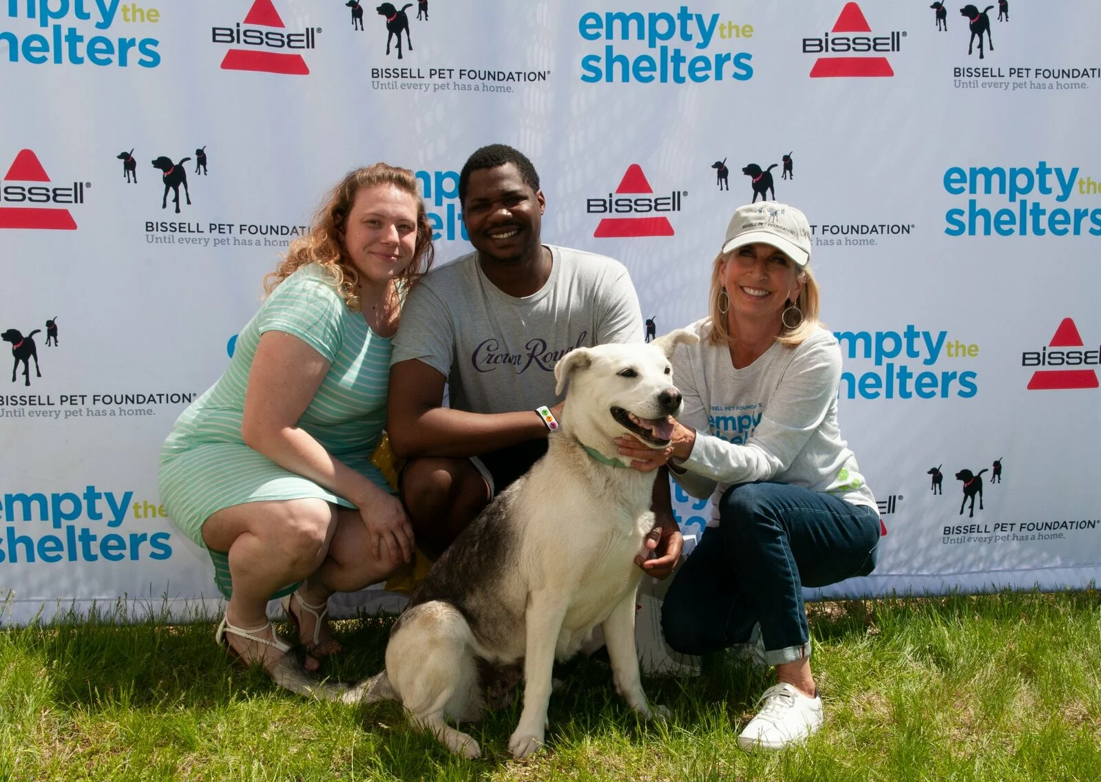 Three people kneel on grass, smiling and posing with a happy husky mix dog in front of a backdrop displaying “empty the shelters” and various sponsor logos.