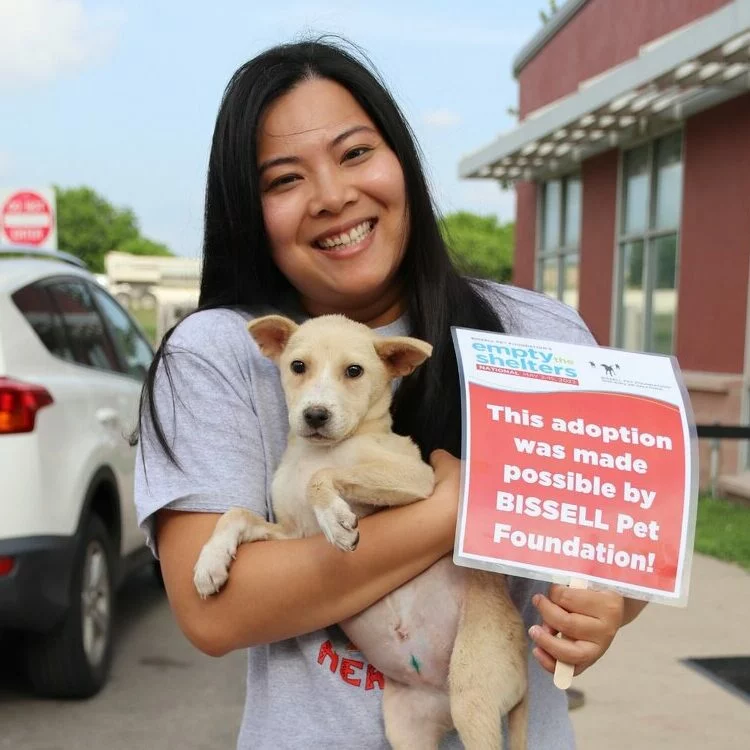 A smiling woman holds a small dog and a sign reading, This adoption was made possible by BISSELL Pet Foundation! outside an animal shelter on a sunny day, celebrating the joy of pet adoption.