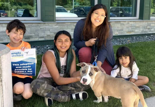 Three children and an adult smile while crouching on grass outside, posing with a light brown and white dog on a leash. One child holds a sign promoting a local pet adoption event.