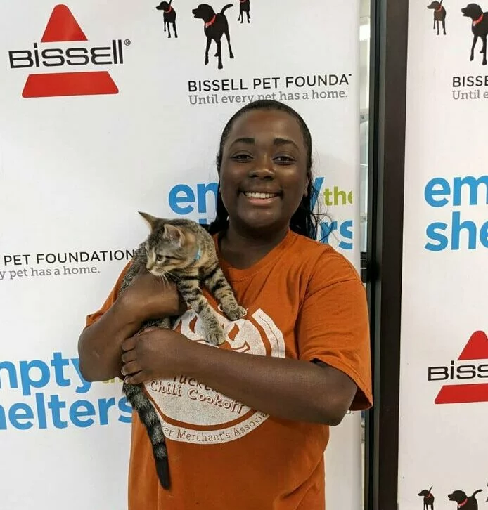 A smiling person in an orange shirt holds a cat in front of a backdrop featuring Empty the Shelters, BISSELL Pet Foundation, and BISSELL logos.