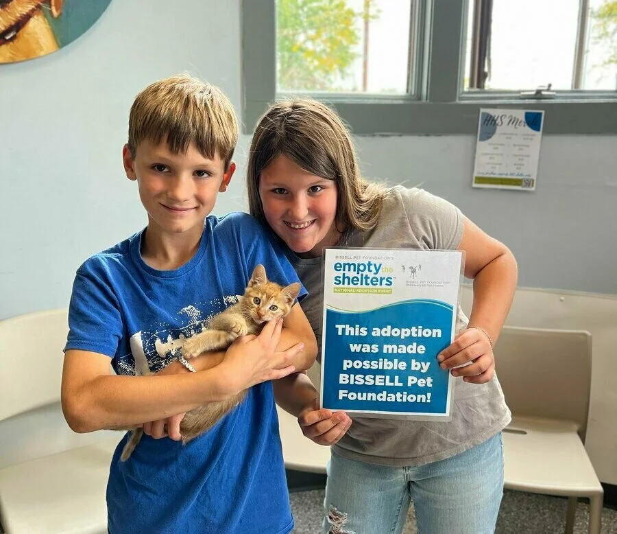Two smiling children stand in an animal shelter waiting area. The boy holds a small tan kitten, while the girl holds a sign reading, “This adoption was made possible by BISSELL Pet Foundation.” 0.