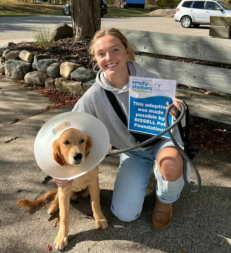 A smiling woman kneels outdoors beside a golden retriever puppy wearing a cone, holding an Empty the Shelters adoption sign. Trees, a wooden bench, and parked cars are in the background on this sunny day.
