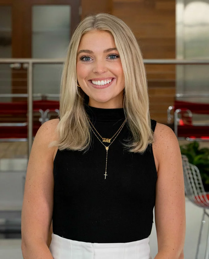 A smiling woman with long blonde hair wears a black sleeveless top, white pants, and gold necklaces. She is standing indoors in a modern setting with wooden and metal accents in the background.