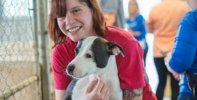 A smiling person in a red shirt hugs a black and white dog inside an animal shelter, with other people blurred in the background.