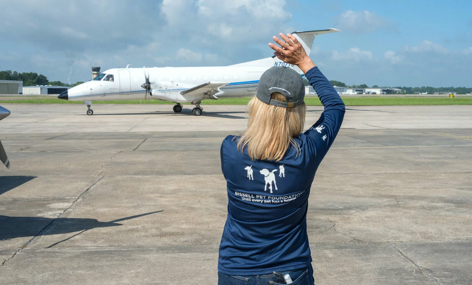 A person in a blue “Bissell Pet Foundation” shirt and cap waves toward a white airplane on an airport tarmac under a partly cloudy sky.