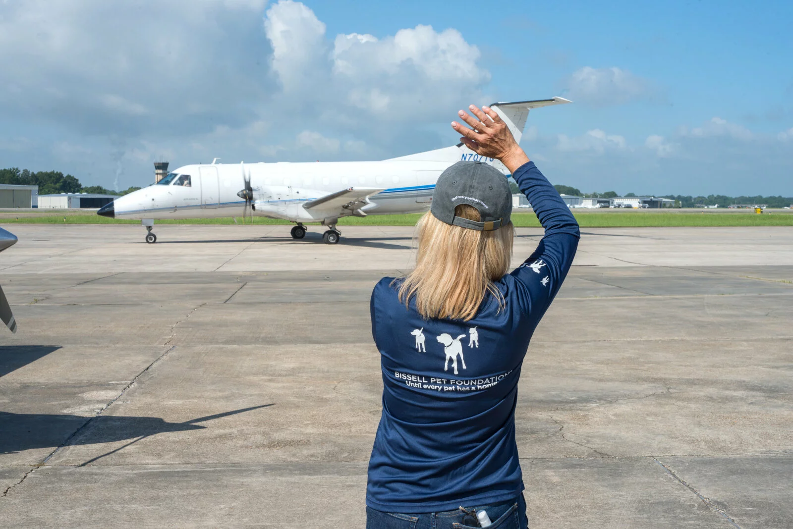 A person wearing a Brishall Pet Foundation shirt stands on an airport tarmac, raising their arms to signal as a small white jet taxiing approaches on a sunny day with blue sky and scattered clouds.