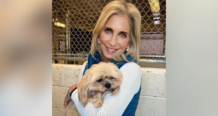A smiling woman with long blonde hair, wearing a white top and blue vest, hugs a small, fluffy dog in front of a chain-link fence and white brick wall indoors, highlighting the importance of vaccines for pets’ health and happiness.