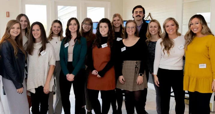 A group of thirteen people, mostly women and one man, stand together indoors, smiling at the camera. Some wear name tags and business casual attire. Bright natural light comes in from windows behind them.