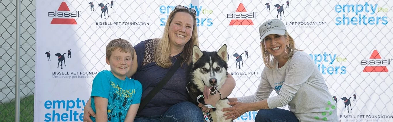 A smiling boy, woman, and another woman pose with a happy husky dog in front of a fence with empty the shelters logos at an outdoor pet adoption event.