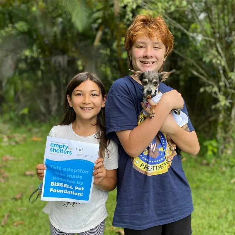 Two smiling children stand outdoors; one holds a small dog and the other proudly displays a sign saying, “This adoption was made possible by BISSELL Pet Foundation!” Lush green trees and grass fill the background.